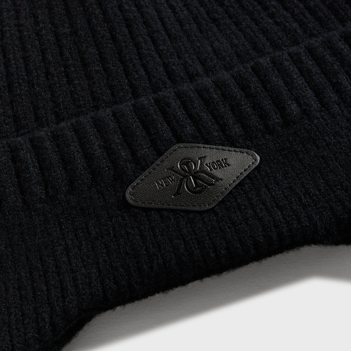 Close-up of the black leather 'New York' diamond patch on the chunky knit earflap beanie